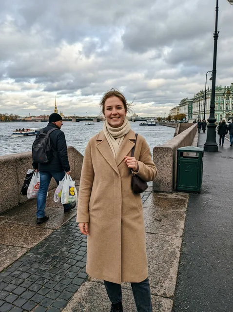 Фото на телефон: девушка в бежевом пальто стоит на набережной Санкт-Петербурга. За ней в кадре прохожий с пакетами и му…